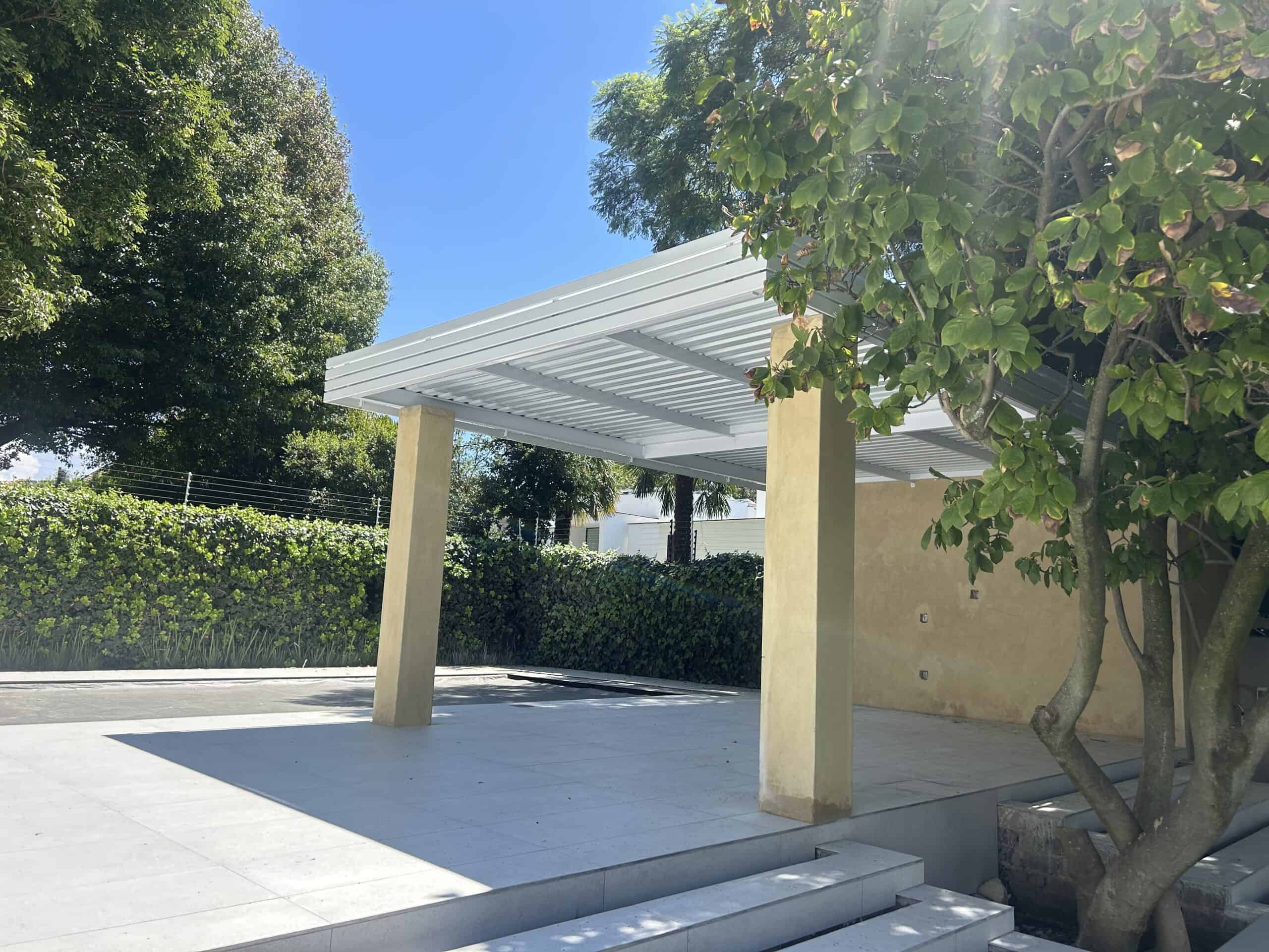Modern outdoor pergola with white slatted roof and beige support columns, surrounded by lush greenery.